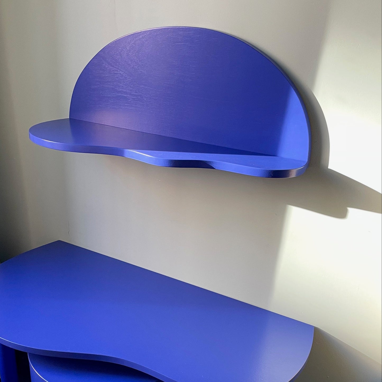 A bright blue children's wall-mounted desk and shelf set with a matching cylindrical stool, shown in a nursery corner next to a crib and patterned curtain.