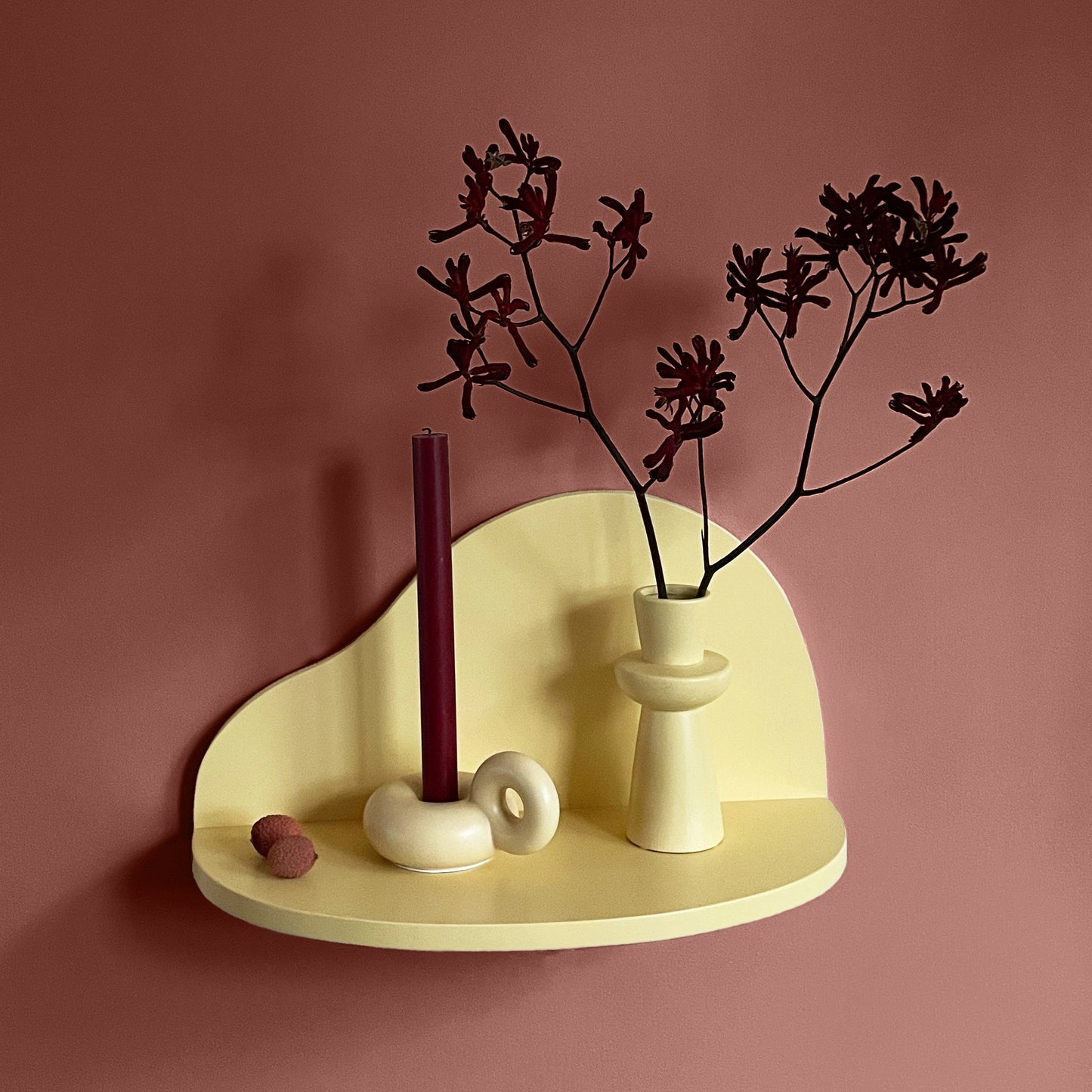 A minimalist cream-colored floating shelf mounted on a dusty rose wall, styled with a dark burgundy candle, a matching vase with dried dark branches, and small decorative stones.