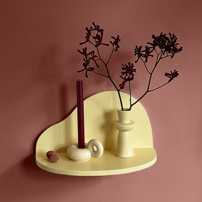A minimalist cream-colored floating shelf mounted on a dusty rose wall, styled with a dark burgundy candle, a matching vase with dried dark branches, and small decorative stones.