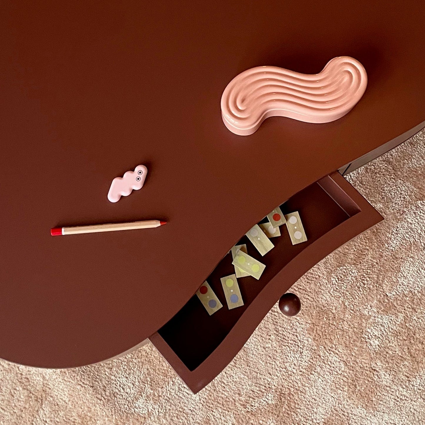 A curved dark brown wooden table with an open drawer, styled with a pink wavy object, a small pink item, and a pencil on a beige textured rug.