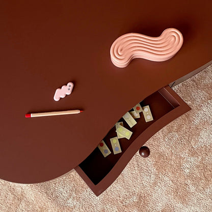 A curved dark brown wooden table with an open drawer, styled with a pink wavy object, a small pink item, and a pencil on a beige textured rug.