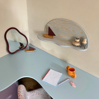 A children's corner desk setup in a nursery or playroom, featuring a wavy light blue desk, a matching curved wall shelf with small wooden toys, a pink-and-white patterned chair, and a small cloud-shaped mirror.