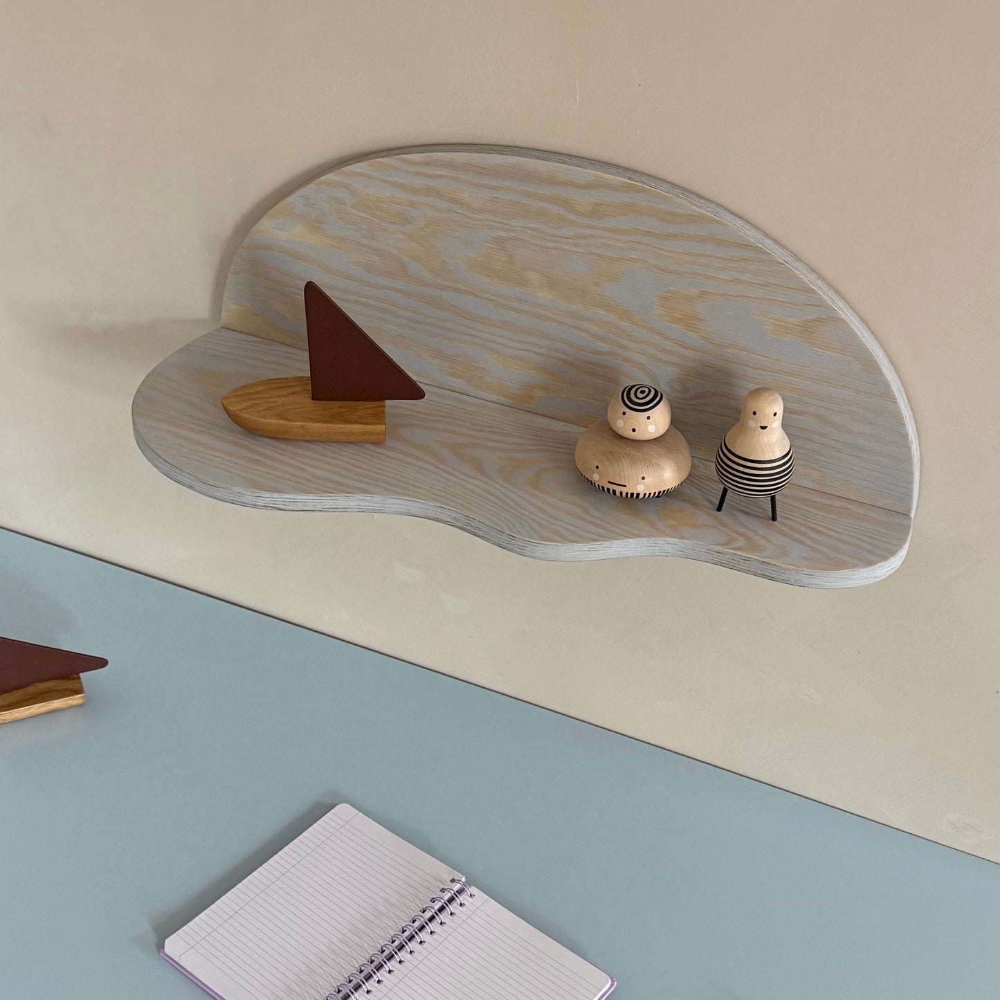 A light gray wood cloud-shaped wall shelf mounted on a beige wall, styled with small wooden toys. Below the shelf is a dusty blue desk surface with a notebook and pencil.