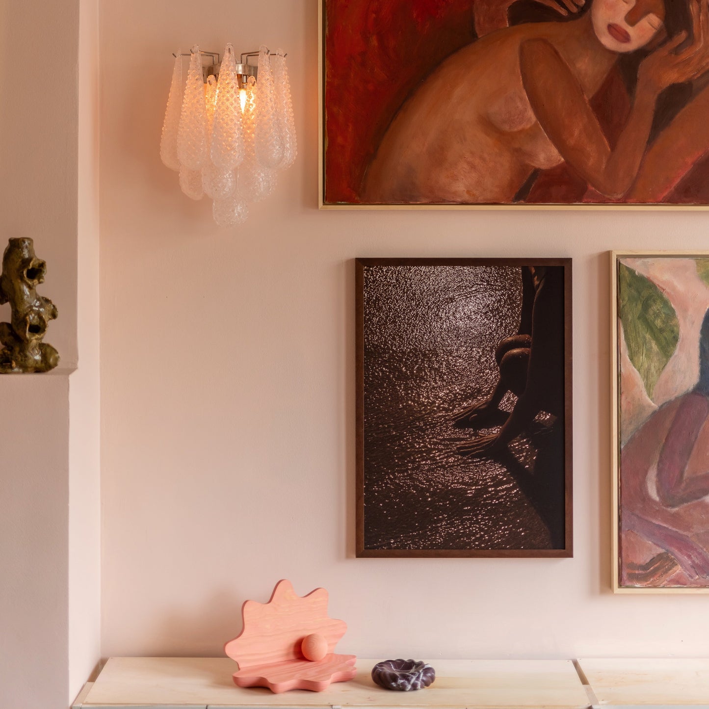 A styled interior wall featuring multiple framed artworks, including a prominent red painting with faces, a dark textured portrait, and a pastel abstract print. A translucent glass wall sconce glows on the left, above a light blue cabinet with pink decorative objects on top.