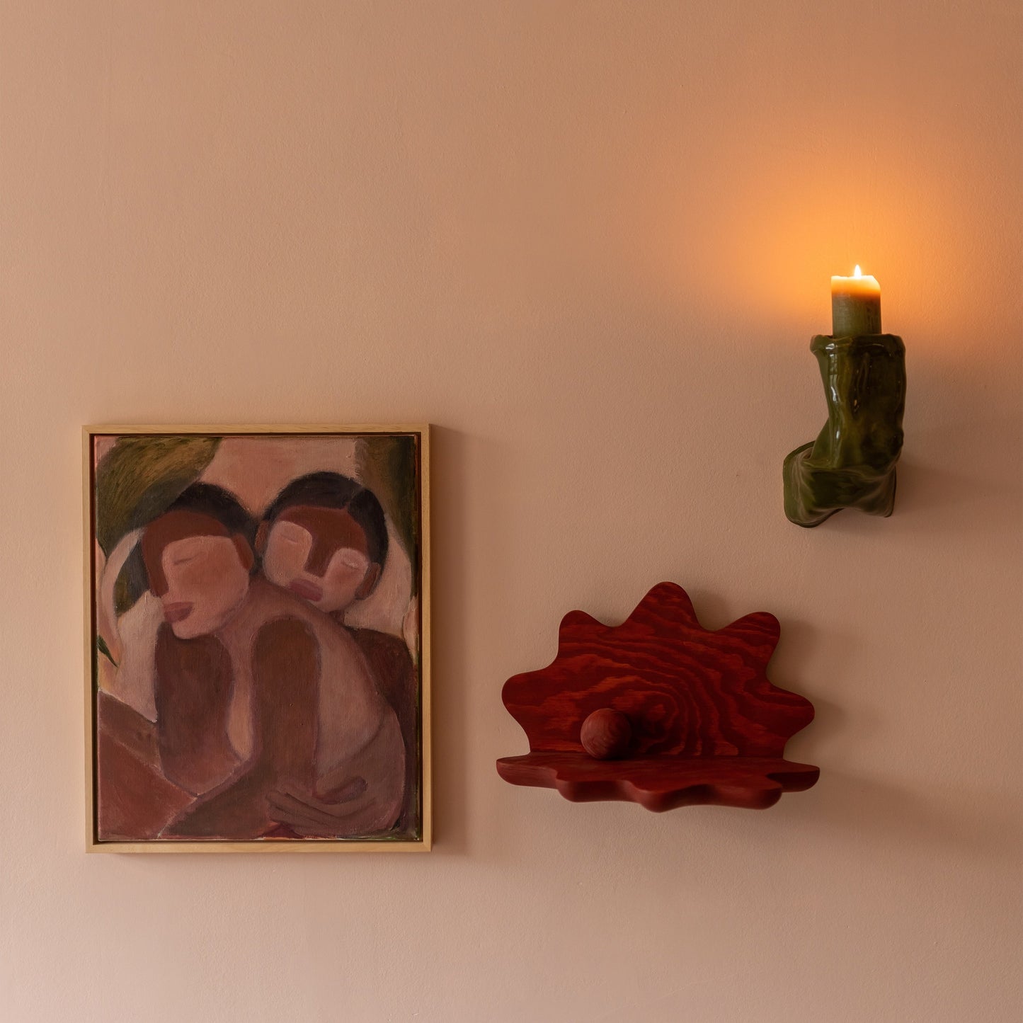 A warm, minimalist interior scene featuring a framed painting of two figures on the left, a red clam-shell shaped wall shelf in the center, and a green sculptural wall sconce holding a lit candle on the right against a soft beige wall.