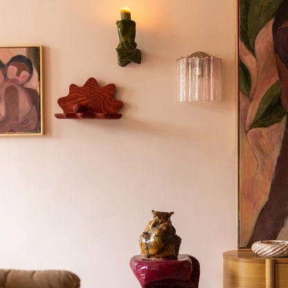A styled interior scene with a glossy magenta sculptural side table holding a brown ceramic vase, set against a warm beige wall with wall art, a green wall sconce, and a clear glass wall light.