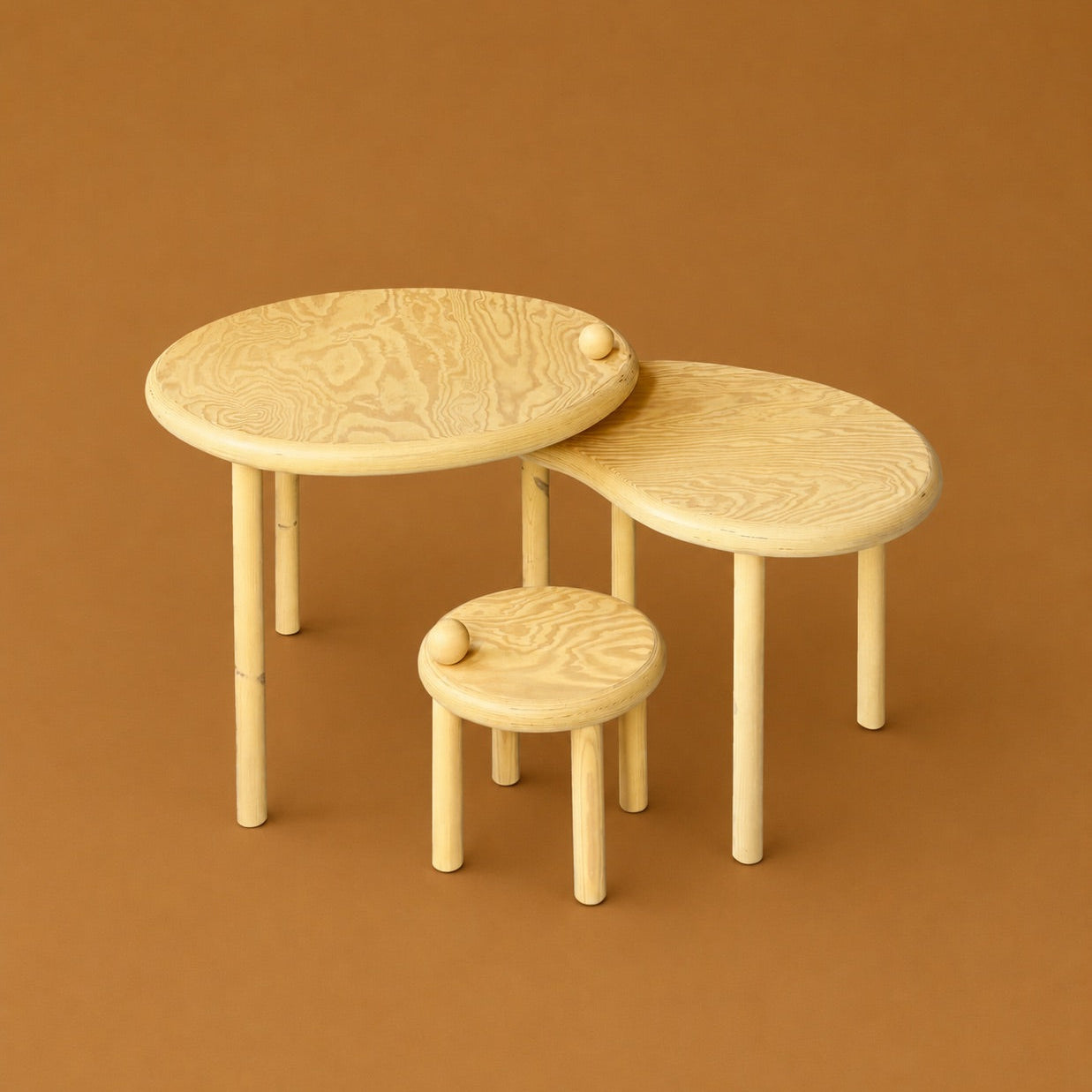 Three light natural wood nesting tables of different oval shapes with matching small stools, set against a solid warm brown background.