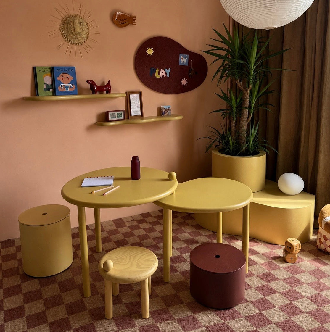 A cozy children's playroom corner with pastel yellow furniture, including a small table, stool, stools, and storage bench, set against a peach-colored wall with shelves, a sun decor piece, and a potted plant.
