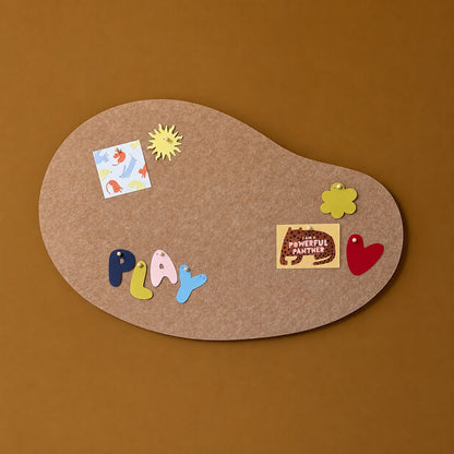 An organic-shaped cork board with a brown textured surface, decorated with colorful paper cutouts including the word 'PLAY', a yellow sun, a flower, a red heart, and a small card reading 'POWERFUL PANTHER'. The board is photographed from above on a solid warm brown background.
