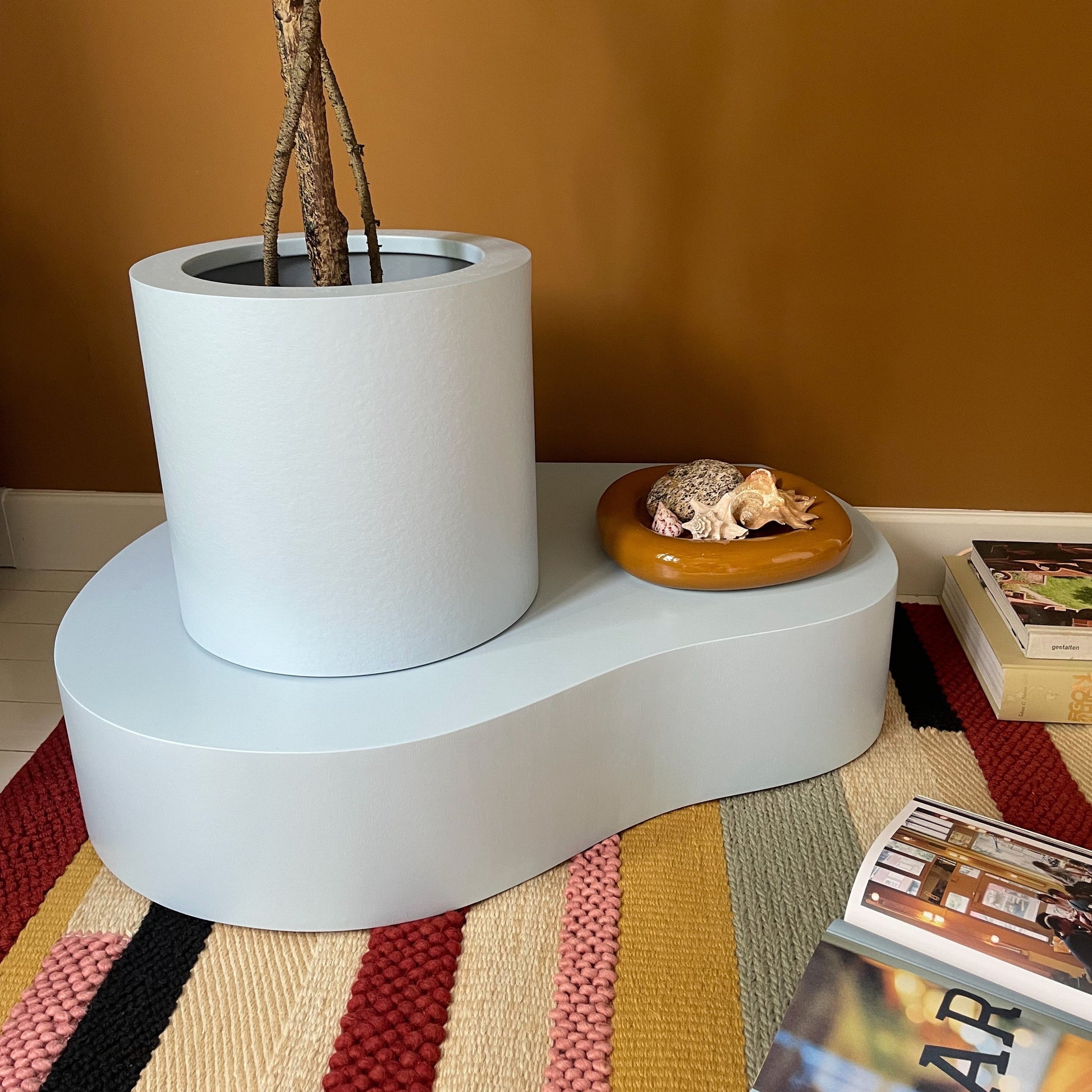 A modern wavy two-tier pedestal in a matte pale blue-gray finish, styled with a large white cylindrical planter holding a tree trunk and a small round wooden tray with a seashell, set on a striped woven rug against a warm mustard wall.