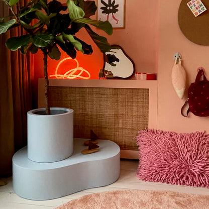 A large potted fiddle-leaf fig plant in a light gray concrete planter displayed on a matching curved pedestal in a cozy, boho-style room with peach walls, a woven cabinet, and a pink shag rug.
