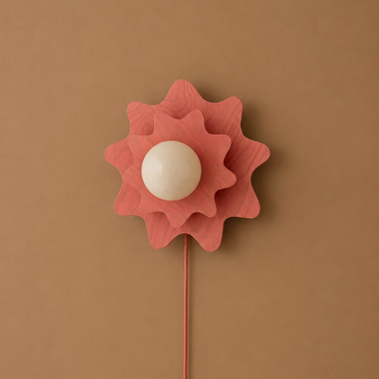 A sculptural flower-shaped wall light or decor piece with layered coral-pink petals, a smooth off-white spherical center, and a thin matching stem against a solid warm tan background.