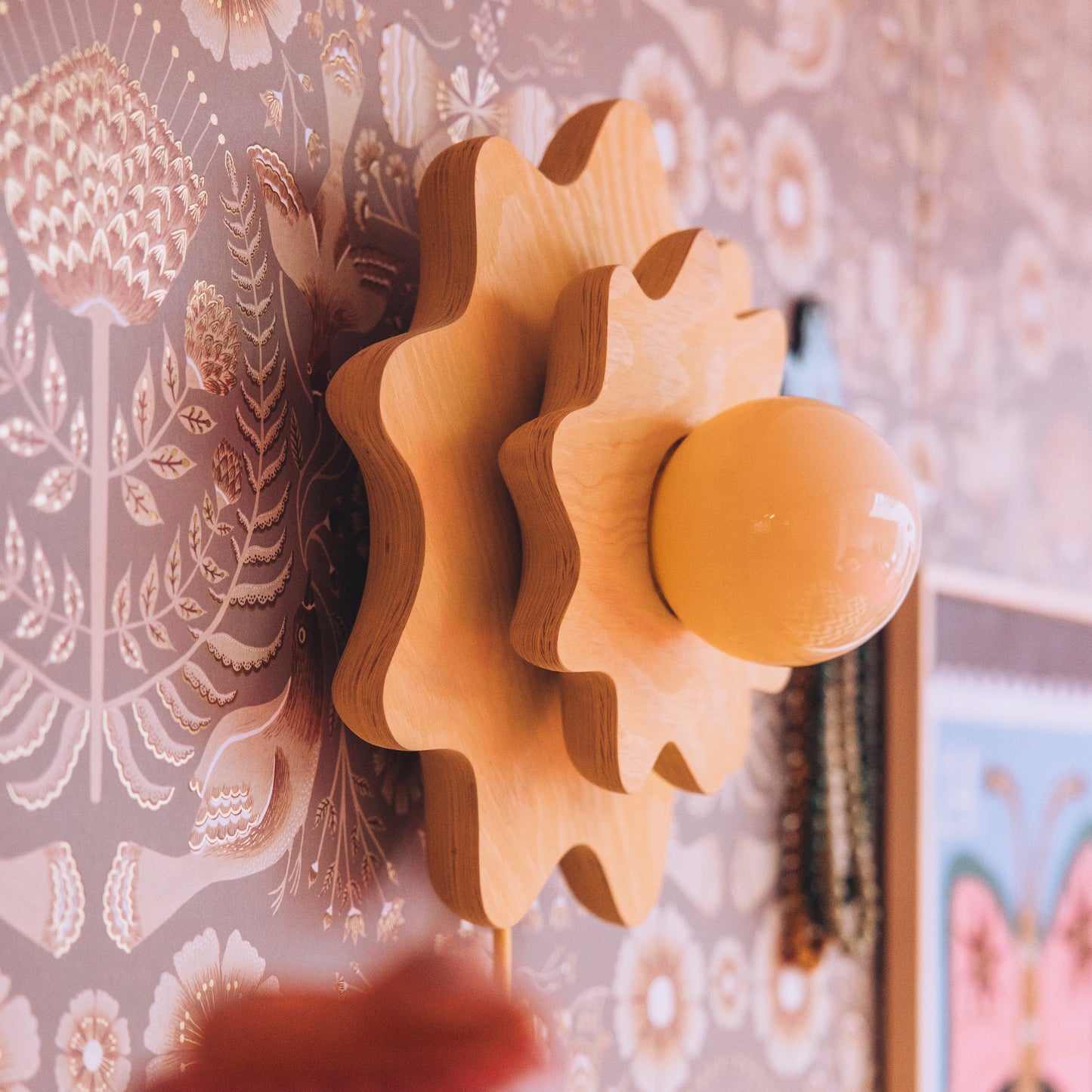 A close-up of a decorative wooden wall sconce with a flower-like layered frame and a round amber globe bulb, mounted against patterned wallpaper.