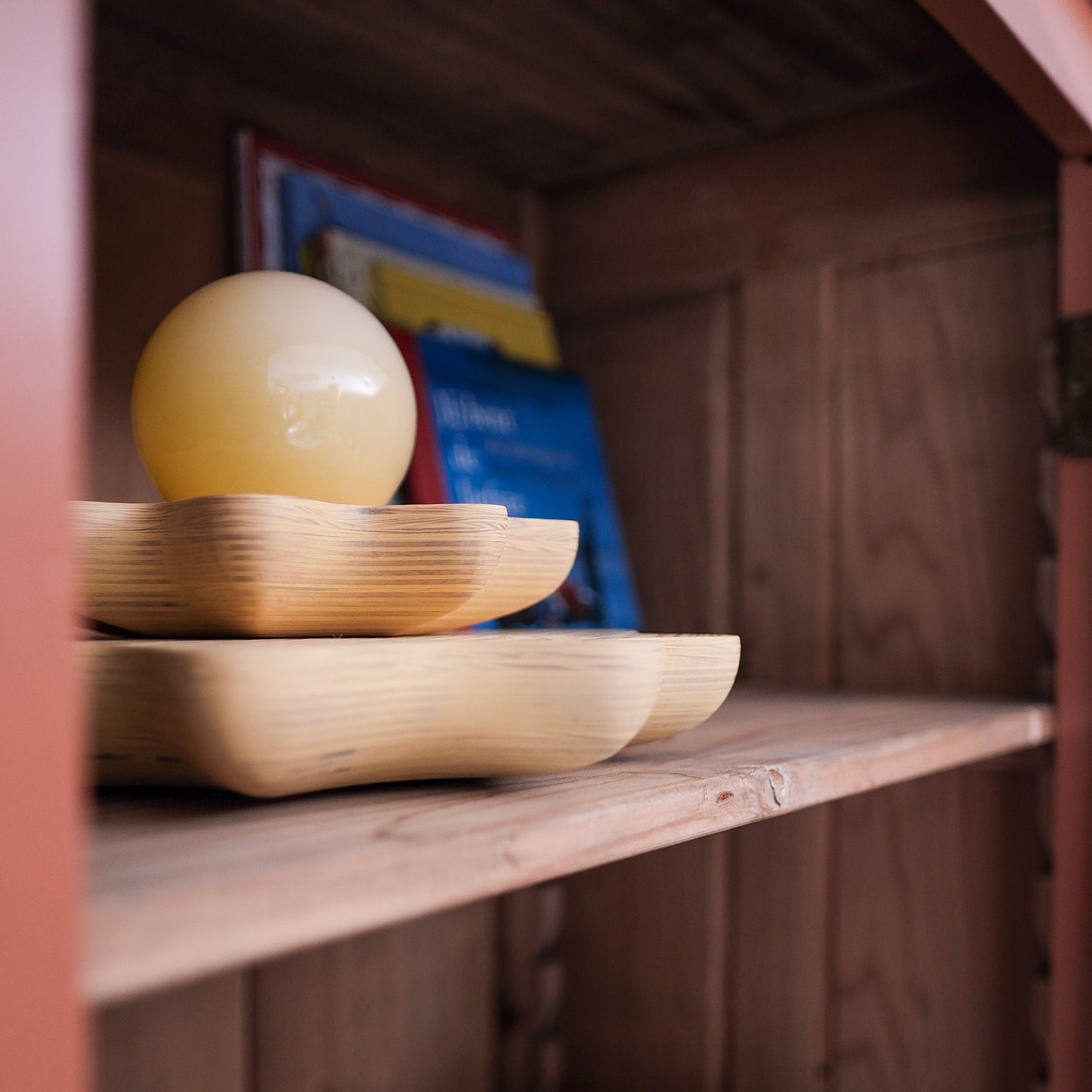 A close-up of a wooden shelf holding a pale yellow oval decorative object on a shallow wooden tray, with books in the background and a purple patterned cushion below.