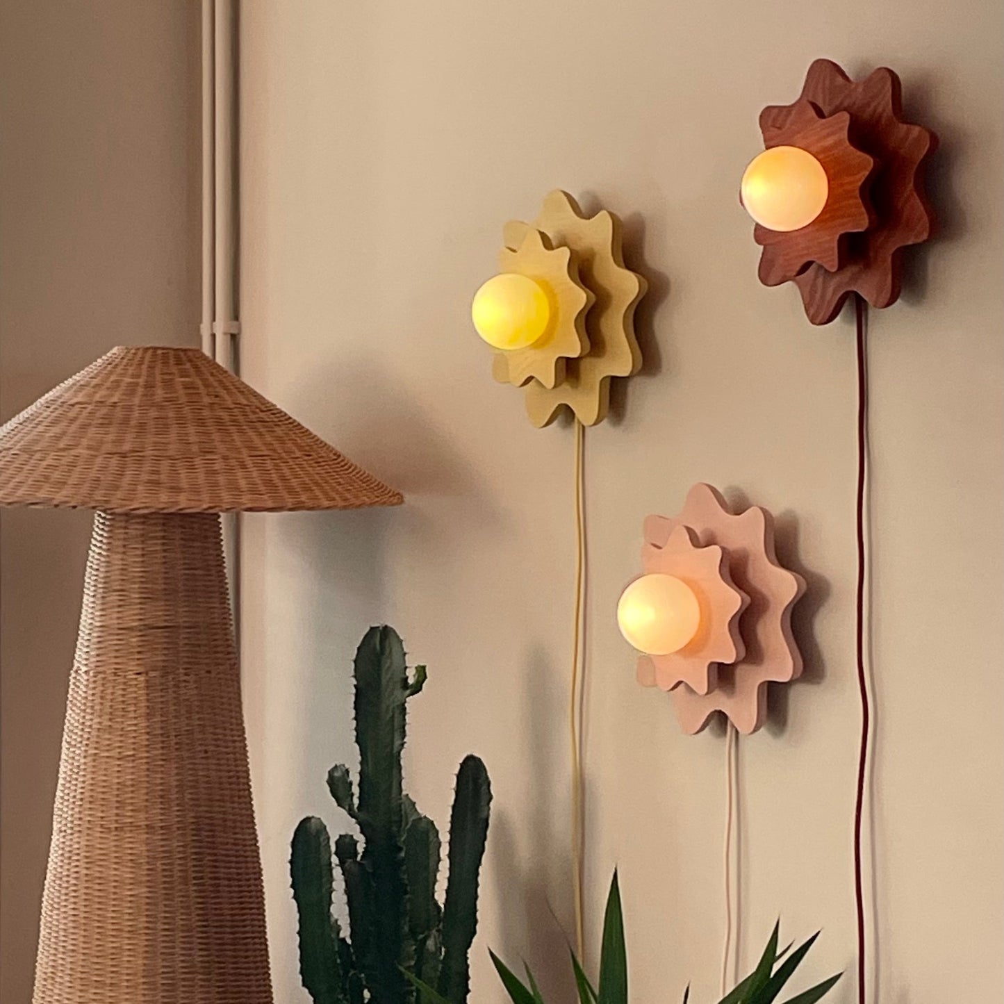 A styled interior scene with a tall woven floor lamp, potted cactus and green plant, and three flower-shaped wall lights in yellow, pink, and brown mounted on a beige wall.