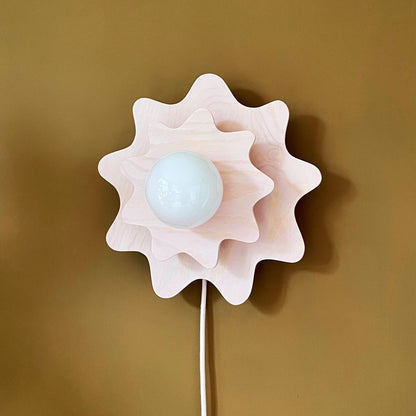 A modern flower-shaped wall lamp in pale pink and white, featuring a central frosted white globe bulb, mounted on a solid mustard yellow wall.