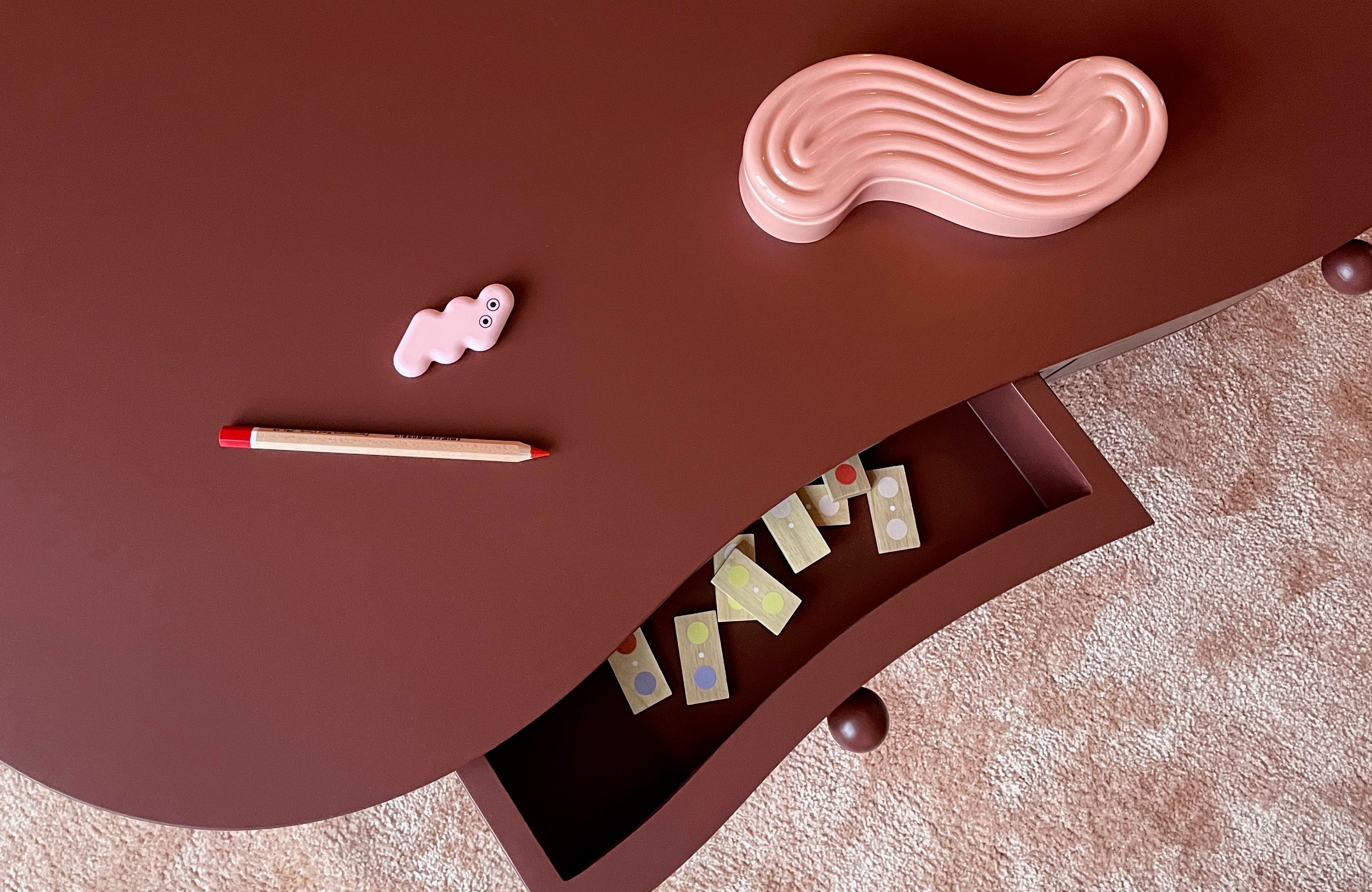 A curved dark brown wooden table with an open drawer, styled with a pink wavy object, a small pink cloud-shaped item, and a wooden pencil on a beige textured rug.