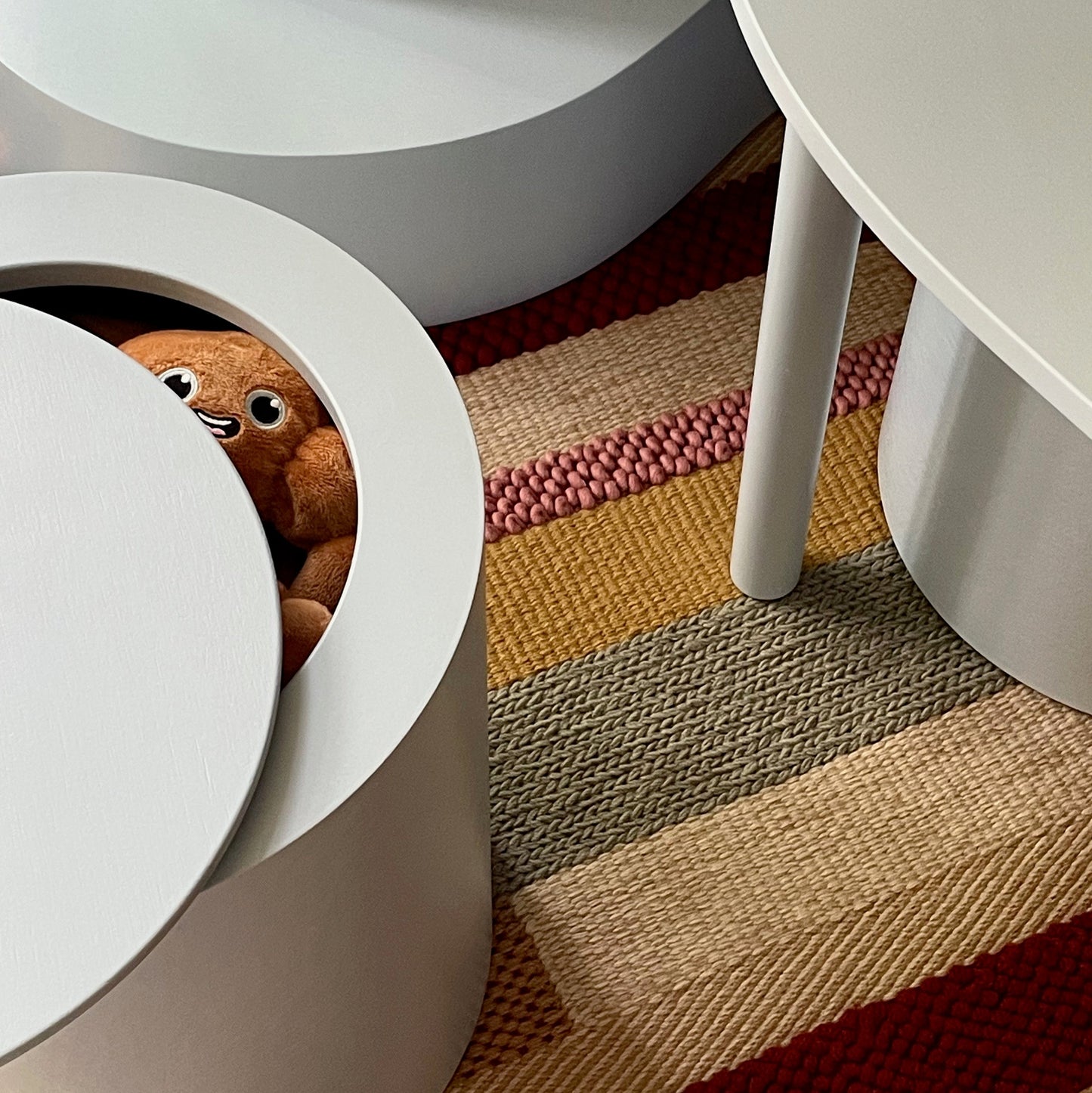 A close-up of a white circular stool or ottoman with a hinged lid, revealing a brown plush toy peeking out. The stool is placed on a striped woven rug with red, beige, and brown tones, next to a white table leg.