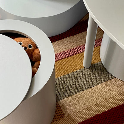 A close-up of a white circular stool or ottoman with a hinged lid, revealing a brown plush toy peeking out. The stool is placed on a striped woven rug with red, beige, and brown tones, next to a white table leg.