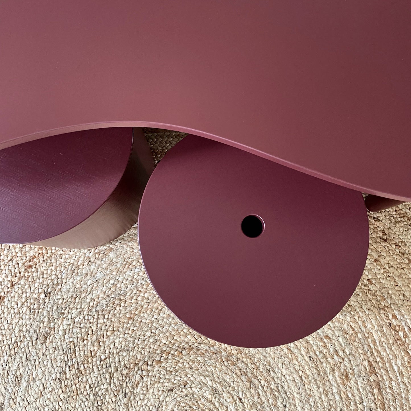 A close-up of a modern, wavy table or stool with a dusty rose pink finish, resting on a textured beige woven rug.