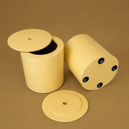 A pale yellow cylindrical container with a matching round lid, shown in two positions on a solid brown background: one standing upright with the lid off, and one lying on its side showing round black fasteners. A separate lid is placed in front.