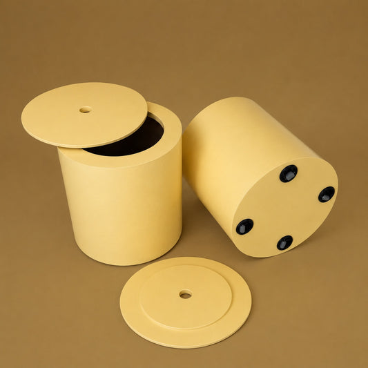A pale yellow cylindrical container with a matching round lid, shown in two positions on a solid brown background: one standing upright with the lid off, and one lying on its side showing round black fasteners. A separate lid is placed in front.