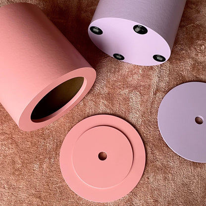 A set of cylindrical and circular components in soft pink and pale lavender, arranged on a textured brown fabric background. One pink cylinder is open, showing the dark interior, while another pale lavender cylinder is shown from the bottom with small black circular details.