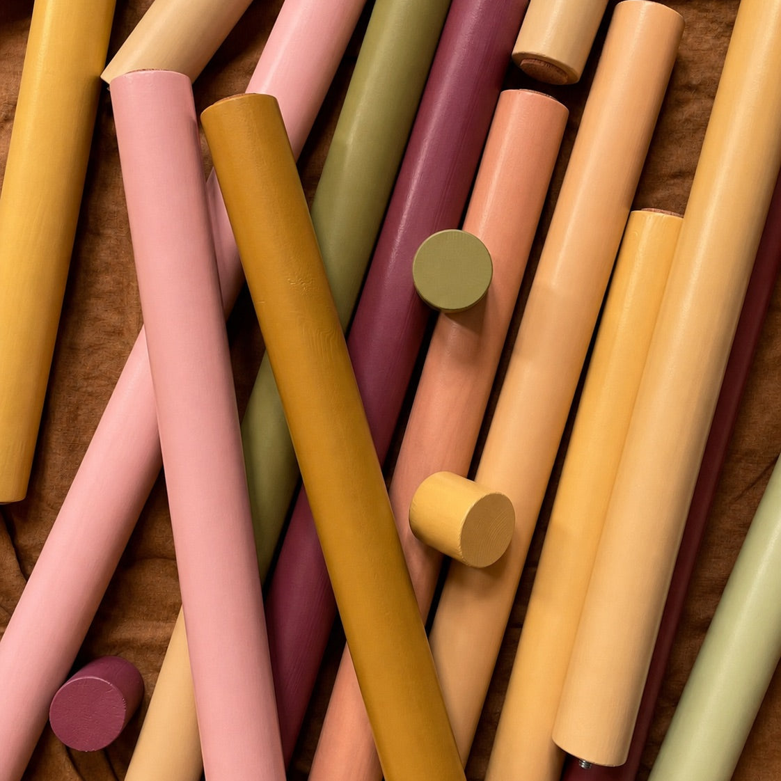 A close-up arrangement of pastel-colored cylindrical rods or straws in pink, mustard yellow, peach, olive green, and mauve, resting on a brown textured fabric background.