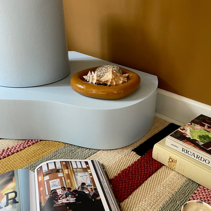 A styled interior scene with a light blue sculptural side table holding a small wooden bowl, next to a white cylindrical lamp base. An open magazine and a book titled RICARDO lie on a striped woven rug, with a small cup nearby against a mustard wall.